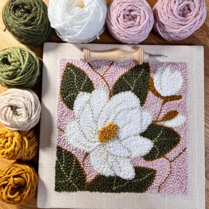 May include: A handmade punch needle art piece featuring a white flower with a yellow center and green leaves on a pink background. Various yarn skeins in green, white, and pink surround the artwork, along with a wooden punch needle tool.