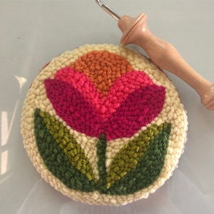 May include: A close-up of a hand-stitched floral design on a round, cream-colored canvas. The design features a pink and orange flower with green leaves. A wooden needle tool is visible in the background.