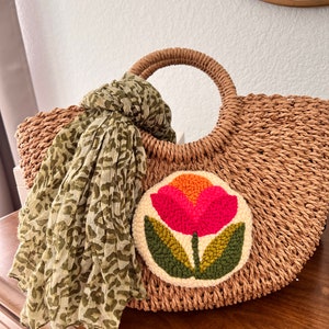 May include: A straw tote bag with a large, round, embroidered flower patch in shades of orange, pink, and green. The bag has a single, large, round handle and a green and white patterned scarf draped over it.