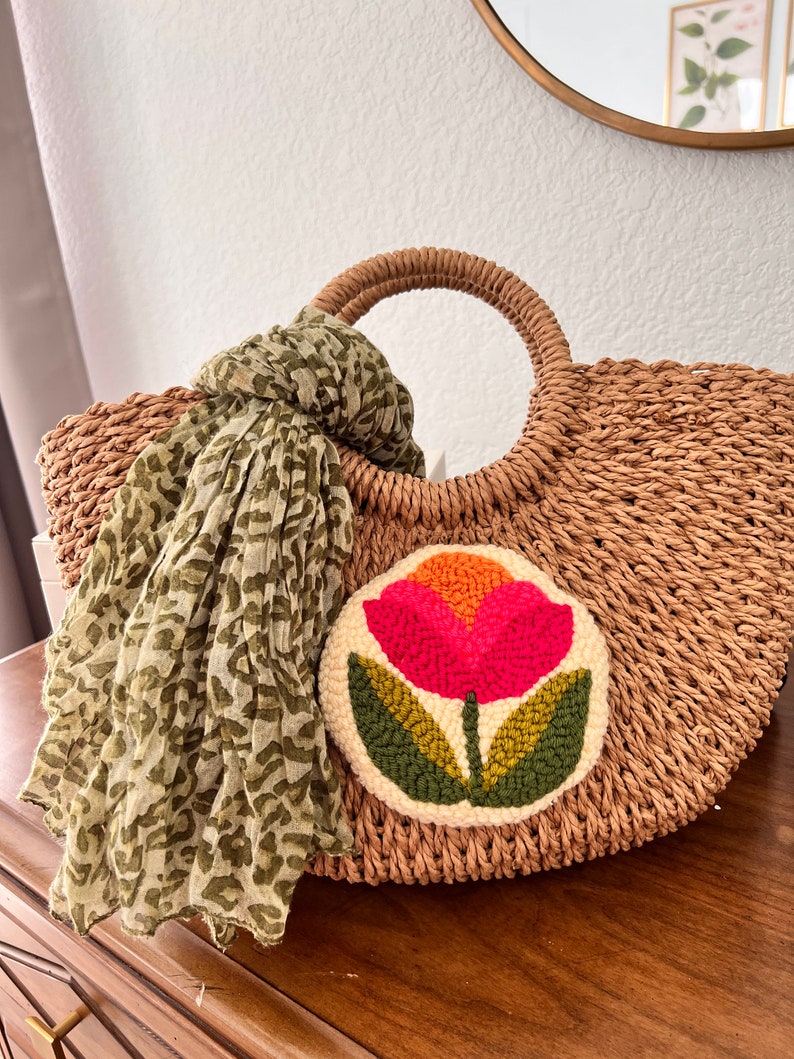 May include: A straw tote bag with a large, round, embroidered flower patch in shades of orange, pink, and green. The bag has a single, large, round handle and a green and white patterned scarf draped over it.