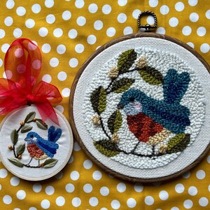 May include: Two embroidered bird hoops. The larger hoop features a blue, red, and beige bird with green leaves. The smaller hoop has a similar design, with a red bow attached. Both are on a yellow polka dot background.