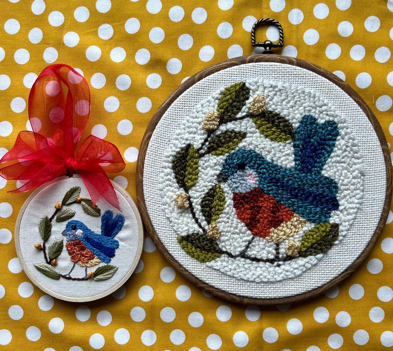 May include: Two embroidered bird hoops. The larger hoop features a blue, red, and beige bird with green leaves. The smaller hoop has a similar design, with a red bow attached. Both are on a yellow polka dot background.