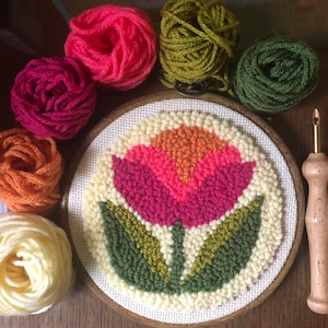 May include: A close-up of a completed embroidery hoop with a colorful flower design. The hoop is surrounded by balls of yarn in various colors, including pink, green, orange, and white.