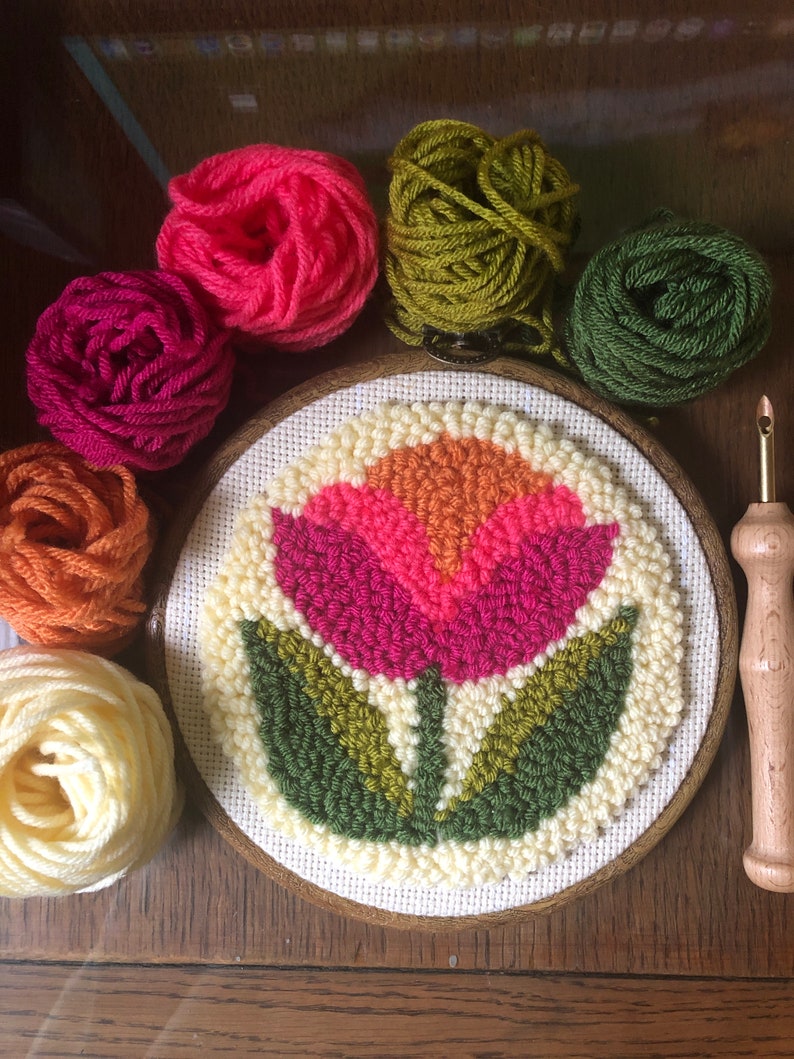 May include: A close-up of a completed embroidery hoop with a colorful flower design. The hoop is surrounded by balls of yarn in various colors, including pink, green, orange, and white.