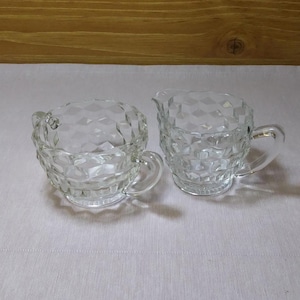 Puede incluir: Un juego de azucarero y jarra de crema de vidrio transparente con un patrón de diamante. El azucarero tiene dos asas y la jarra de crema tiene una sola.