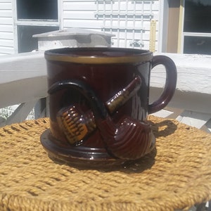 Peut inclure: Une tasse en céramique brune avec un motif de pipe. La tasse a une anse et une soucoupe. La tasse est posée sur un dessous de tasse en osier.