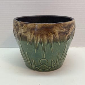 Peut inclure: Un pot de fleurs en céramique avec un intérieur marron foncé et un extérieur bicolore. La partie supérieure est marron, passant à une base vert turquoise avec un motif texturé et sculpté. Le pot a une forme ronde.