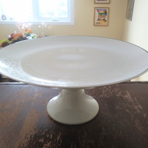 May include: White ceramic cake stand with a silver rim and a delicate floral pattern. The stand has a round base and a single pedestal.