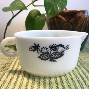 Puede incluir: Jarrita de leche de vidrio blanco con un diseño floral negro. La jarrita tiene una forma redondeada y un asa.