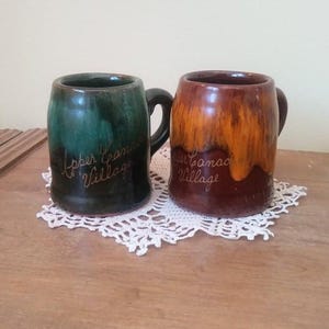 Peut inclure: Deux mugs en céramique avec des anses, un vert et un marron, posés sur un napperon blanc. Le mug vert porte l'inscription "Upper Canada Village". Le mug marron porte l'inscription "Upper Canada Village".