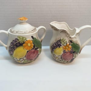 Op de afbeelding: Een crèmekleurige keramische suikerpot met deksel en een melkkan. Beide stukken zijn versierd met een kleurrijk fruit- en bloemmotief, waaronder druiven, citroenen en peren. De suikerpot heeft twee handvatten en een kleine oranje knop.