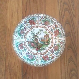 Peut inclure: Une assiette ronde en céramique blanche avec un motif floral. Le centre de l'assiette présente une scène colorée avec un bâtiment, des arbres et un bateau. L'assiette a un bord festonné et est décorée d'une bordure brune et dorée.