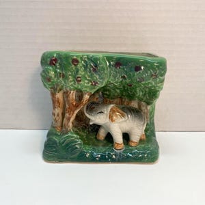 Puede incluir: Una maceta de cerámica con una escena de bosque y una pequeña figura de elefante. La maceta tiene una base verde y árboles con troncos marrones y follaje verde. El elefante es gris, blanco y beige.