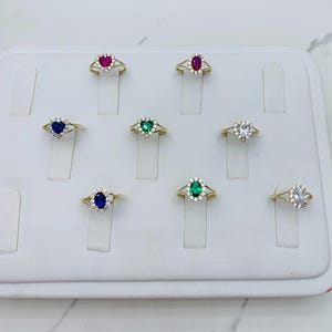 May include: A display of nine gold rings with different colored gemstones. The rings are set on a white display board. The gemstones are red, green, blue, and white.