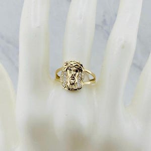 May include: A gold-colored ring featuring a detailed depiction of Jesus' face. The ring has a halo design and is adorned with small, clear gemstones. The ring is displayed on a light-colored surface.