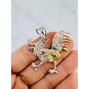 Può includere: Un ciondolo a forma di gallo in metallo tricolore nei toni dell'oro bianco, giallo e rosa. Il gallo presenta un piccolo occhio in pietra preziosa rossa e pietre chiare incastonate lungo le piume dell'ala e della coda, con una contromaglia.