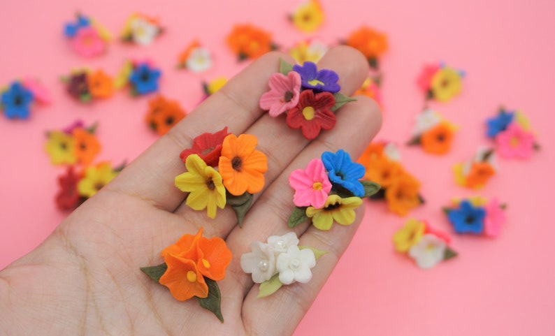 Pode incluir: Uma cole&ccedil;&atilde;o de pequenos aglomerados de flores coloridas em v&aacute;rias tonalidades, incluindo laranja, amarelo, rosa, vermelho e azul. Cada aglomerado apresenta m&uacute;ltiplas p&eacute;talas e folhas verdes, num fundo rosa.