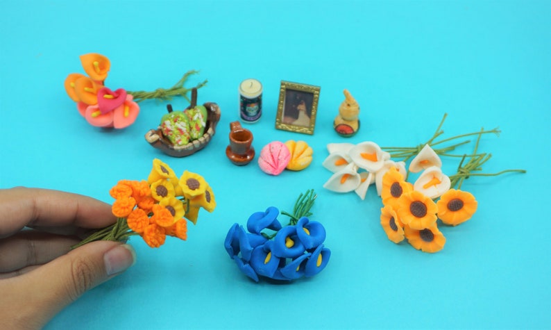 May include: Assortment of miniature artificial flower bouquets in various colors, including orange, yellow, white, blue, and pink. The bouquets are arranged on a blue surface with other miniature items, such as a small picture frame, a ceramic mug, and a small ceramic bowl.