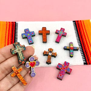 May include: A collection of small, flat, cross-shaped ornaments featuring vibrant, multi-colored floral and heart-themed patterns. Each cross has a black edge and displays distinct designs, including bright orange, pink, blue, and green motifs.