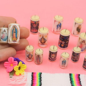 May include: A collection of miniature cylindrical candles featuring various religious and cultural imagery, including skulls, figures, and floral patterns. The candles are white with printed designs and a single black wick on top of each small wax cylinder.