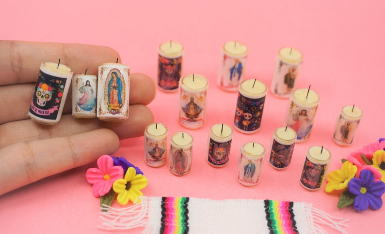 May include: A collection of miniature cylindrical candles featuring various religious and cultural imagery, including skulls, figures, and floral patterns. The candles are white with printed designs and a single black wick on top of each small wax cylinder.