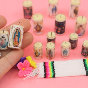May include: A collection of miniature cylindrical candles featuring various religious and cultural imagery, including a sugar skull with the text Dia de Muertos, alongside a small white woven mat with colorful stripes and plastic flower accents.