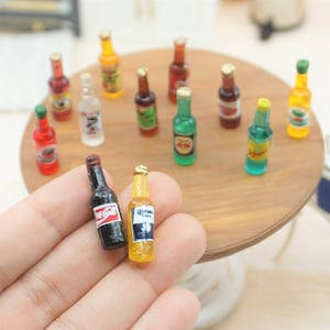 May include: Miniature bottles of various beverages, including Coca-Cola and Corona, are arranged on a wooden table. The bottles are colorful and detailed, with labels and caps. A hand holds two bottles in the foreground. The scene suggests a bar or restaurant setting.