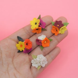 Pode incluir: Uma cole&ccedil;&atilde;o de pequenas flores de papel coloridas em tons de amarelo, laranja, roxo, branco e bord&ocirc;. As flores s&atilde;o dispostas em pequenos grupos com folhas verdes, sobre um fundo rosa.