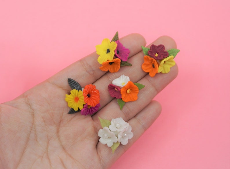 Pode incluir: Uma cole&ccedil;&atilde;o de pequenas flores de papel coloridas em tons de amarelo, laranja, roxo, branco e bord&ocirc;. As flores s&atilde;o dispostas em pequenos grupos com folhas verdes, sobre um fundo rosa.