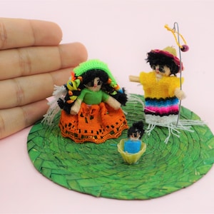 Könnte beinhalten: Ein Set aus drei kleinen handgefertigten Puppen auf einer grünen gewebten Basis. Die Puppen sind in farbenfrohe Kleidung gekleidet und haben schwarzes Haar. Die weibliche Puppe trägt ein grünes Hemd und einen orangefarbenen Rock. Die männliche Puppe trägt ein gelbes Hemd und einen blau-rosa gestreiften Rock. Die Baby-Puppe trägt ein blaues Hemd.