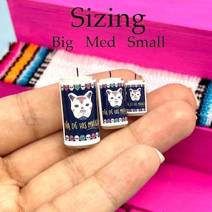 May include: Three cylindrical candles in big, medium, and small sizes, each featuring a dark blue label with a white sugar skull dog face, colorful skull borders, and the text Dia de los Muertos. The candles have a single black wick each.