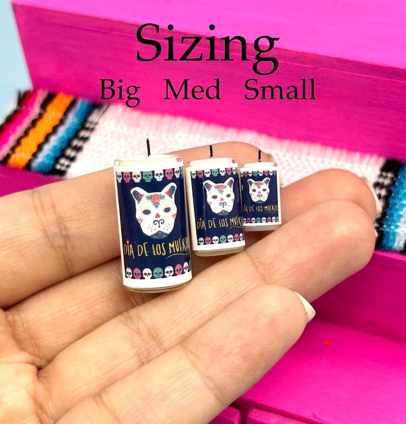 May include: Three cylindrical candles in big, medium, and small sizes, each featuring a dark blue label with a white sugar skull dog face, colorful skull borders, and the text Dia de los Muertos. The candles have a single black wick each.