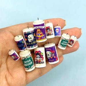 May include: A collection of 10 small white candles with colorful Day of the Dead designs. The candles feature images of skulls, flowers, and the text "Dia de los Muertos" and "Viva Mexico".