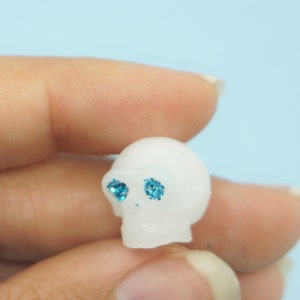Könnte beinhalten: Kleine, weiße Totenkopf-Figur mit blau glitzernden Augen. Der Schädel wird zwischen Fingern vor einem hellblauen Hintergrund gehalten. Dieser Miniatur-Schädel ist ein dekorativer Artikel, möglicherweise für Halloween oder Gothic-Themen.