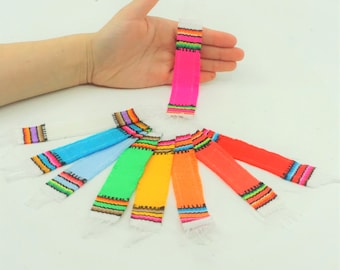 May include: A set of eight colorful mini Mexican blankets with fringe. The blankets are in various colors, including pink, blue, green, yellow, and orange. Each blanket has a white border and a black and white geometric pattern.