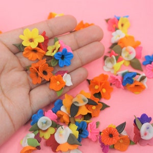 Pode incluir: Uma cole&ccedil;&atilde;o de pequenas flores de tecido coloridas em tons de laranja, amarelo, azul, rosa e branco, com folhas verdes. As flores est&atilde;o espalhadas sobre uma superf&iacute;cie rosa, algumas sendo seguradas em uma m&atilde;o.
