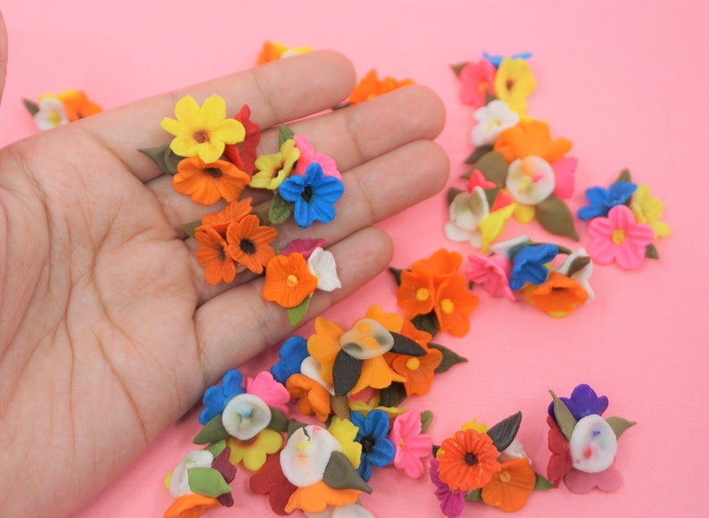 Pode incluir: Uma cole&ccedil;&atilde;o de pequenas flores de tecido coloridas em tons de laranja, amarelo, azul, rosa e branco, com folhas verdes. As flores est&atilde;o espalhadas sobre uma superf&iacute;cie rosa, algumas sendo seguradas em uma m&atilde;o.