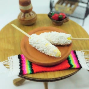 May include: Miniature corn on the cob, coated in a white topping, on a terracotta plate. The plate rests on a small wooden table with a colorful woven table runner. A small wooden chair and other miniature food items are also visible.