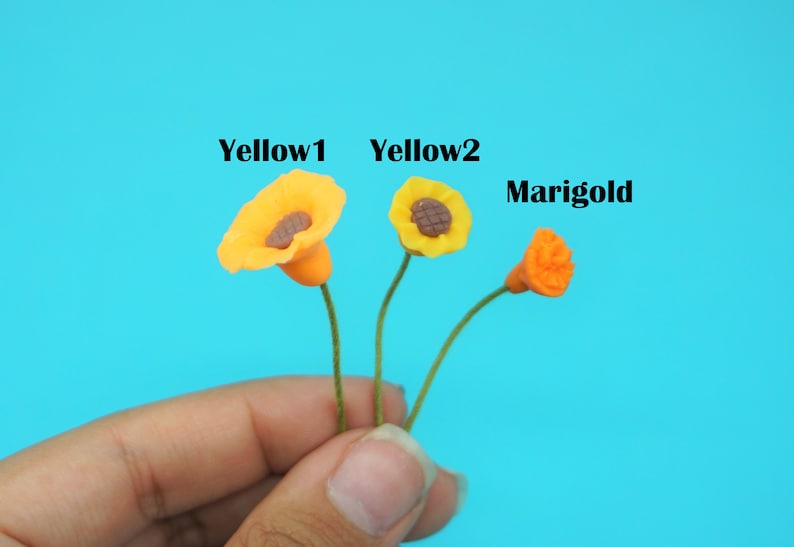 May include: Three miniature clay flowers, labeled "Yellow1", "Yellow2", and "Marigold". The flowers are yellow and orange, with brown centers. The flowers are held in a hand.