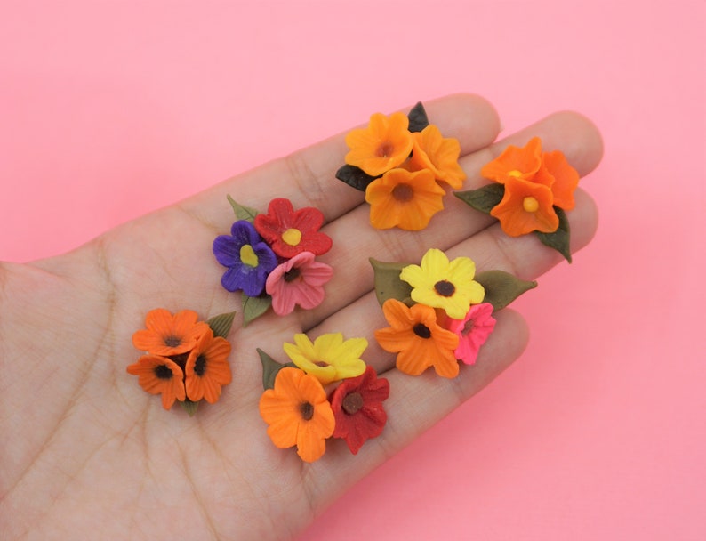 Pode incluir: Um conjunto de cachos de flores em miniatura, coloridos, em tons de laranja, amarelo, vermelho, rosa e roxo. Cada cacho apresenta p&eacute;talas detalhadas e folhas verdes, sobre um fundo rosa suave.