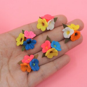 Pode incluir: Uma cole&ccedil;&atilde;o de cachos de flores em miniatura coloridas. Cada cacho apresenta uma mistura de cores vibrantes como rosa, amarelo, laranja, azul e branco, com folhas verdes. As flores est&atilde;o dispostas sobre um fundo rosa.