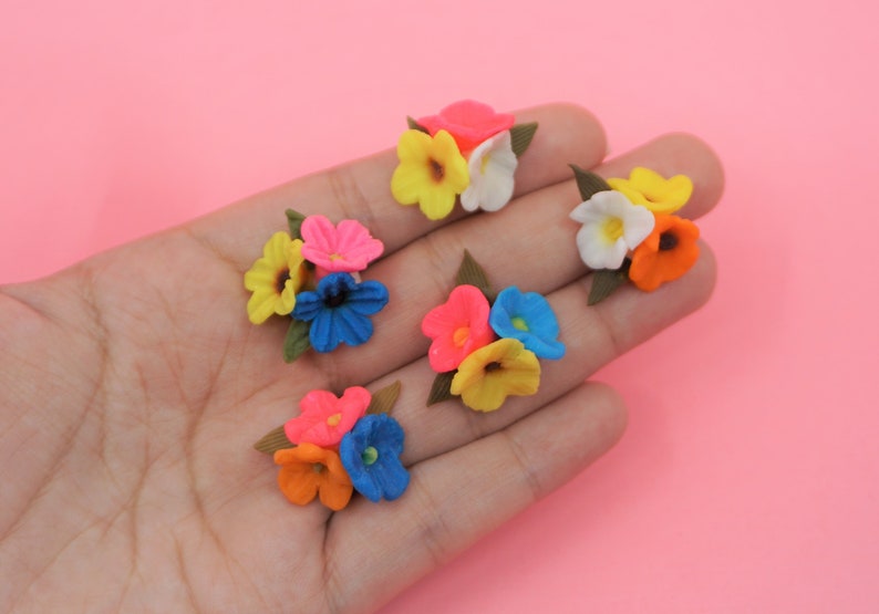 Pode incluir: Uma cole&ccedil;&atilde;o de cachos de flores em miniatura coloridas. Cada cacho apresenta uma mistura de cores vibrantes como rosa, amarelo, laranja, azul e branco, com folhas verdes. As flores est&atilde;o dispostas sobre um fundo rosa.