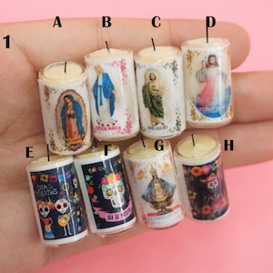 May include: A collection of eight miniature cylindrical candles labeled A through H, featuring religious and cultural imagery including Our Lady of Guadalupe, Virgin Mary, Saint Jude, Jesus, and Dia de los Muertos sugar skulls with floral accents.
