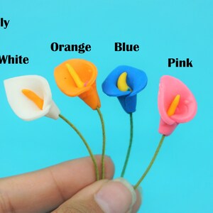 May include: Five artificial Calla Lily flowers in white, orange, blue, and pink. The flowers are held in a hand against a blue background.