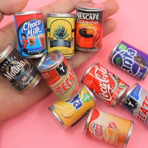 May include: A collection of miniature metal cans with various brand names and designs, including Coca-Cola, Tecate, Modelo, and Nescafe. The cans are arranged on a hand, with a pink background.