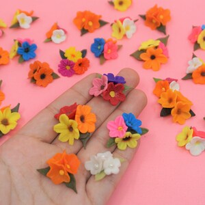 Pode incluir: Uma cole&ccedil;&atilde;o de pequenos bouquets de flores coloridas em tons de laranja, amarelo, rosa, azul e branco. Cada bouquet tem folhas verdes. As flores est&atilde;o espalhadas numa superf&iacute;cie rosa.
