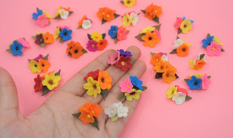Pode incluir: Uma cole&ccedil;&atilde;o de pequenos bouquets de flores coloridas em tons de laranja, amarelo, rosa, azul e branco. Cada bouquet tem folhas verdes. As flores est&atilde;o espalhadas numa superf&iacute;cie rosa.