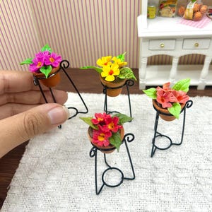 May include: Miniature flower pots with pink, yellow, and red blooms. Each pot is on a black metal stand, creating a charming display. The flowers have green leaves. The background is white.