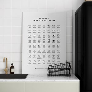 Może przedstawiać: Czarno-biały plakat z tekstem "Laundry Care Symbol Guide" i siatką symboli pielęgnacji odzieży. Plakat wisi na białej, kafelkowej ścianie nad kuchennym zlewem.