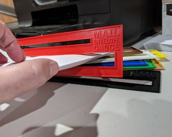 May include: Red plastic mail envelope gauge with the text "MAIL ENVELOPE GAUGE" printed on it. A white envelope is being inserted into the gauge.